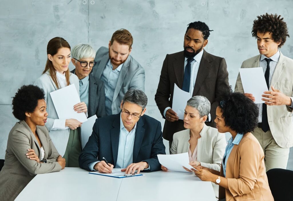 Group of people in business attire all looking at the person in the center who is signing a document. Topic: hiring digital freelancers