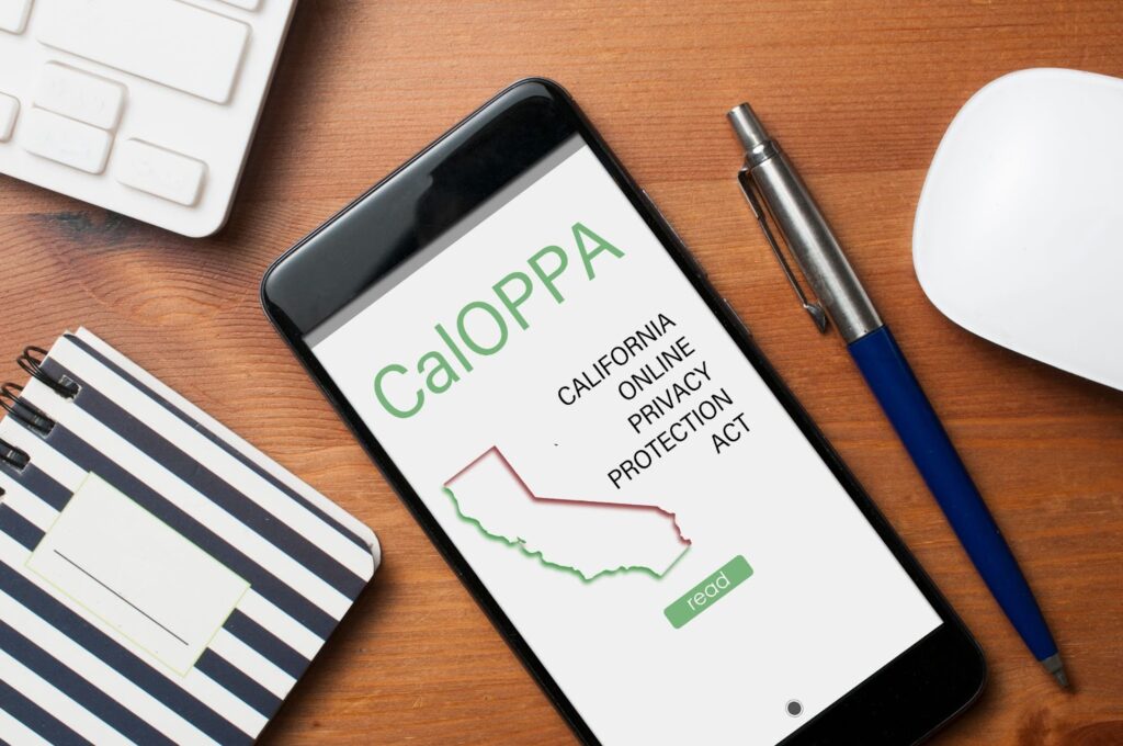 Phone face up on desk with screen that reads "CalOPPA California Online Privacy Protection Act, read". Surrounding the phone is a corner image of the computer keyboard, a notepad, and pen. Topic: California Privacy Policy