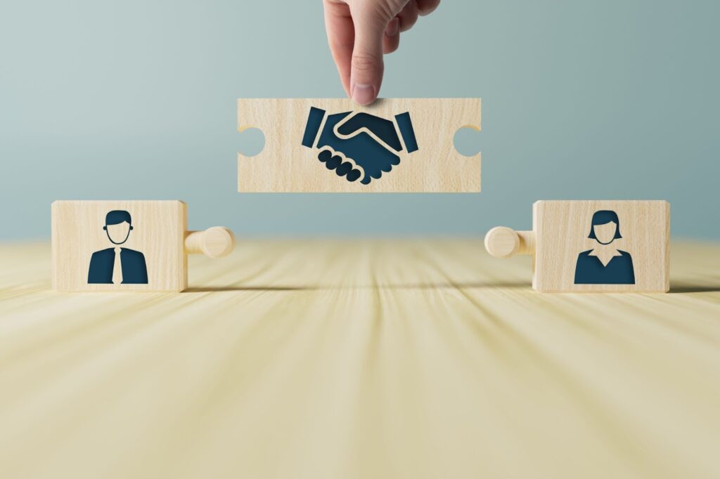 Image of 3 wooden puzzle pieces. One piece on the left shows an image of a man with image on the right shows an image of a woman. Connecting the two images in the middle shows a handshake. Topic: Arbitration Clause in Online Course Terms
