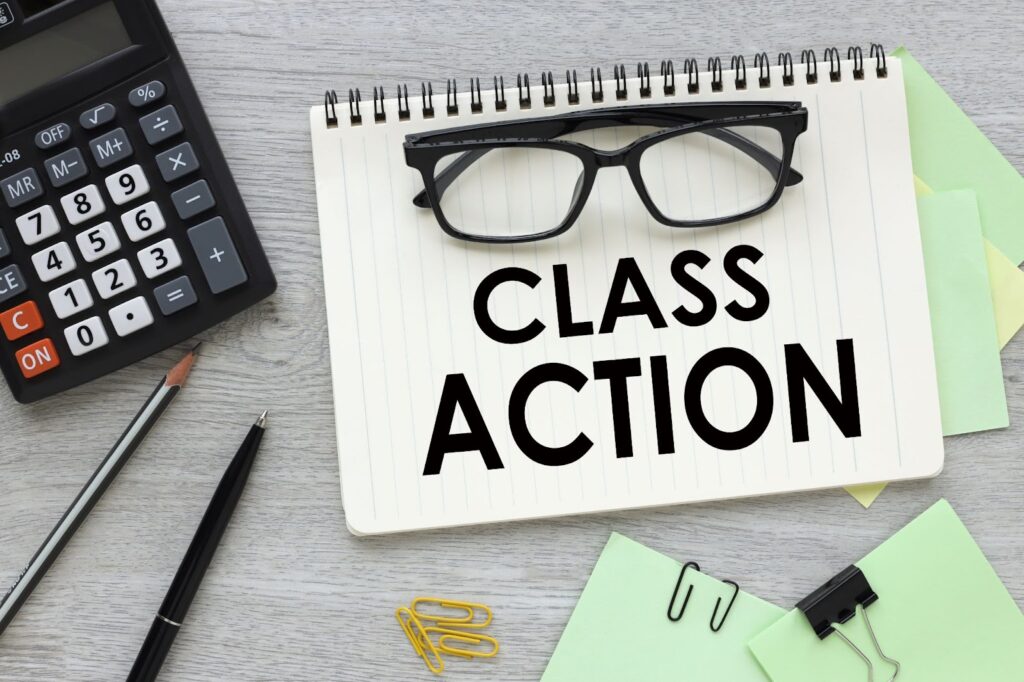 The phrase "Class Action" is written in bold font on a notebook page. Eyeglasses folded and placed on top of the book above the phrase. Notebook is placed on a desk that also has a calculator, pencil, pen, paperclip and scrap paper scattered around. Topic: Arbitration Clause in Online Course Terms